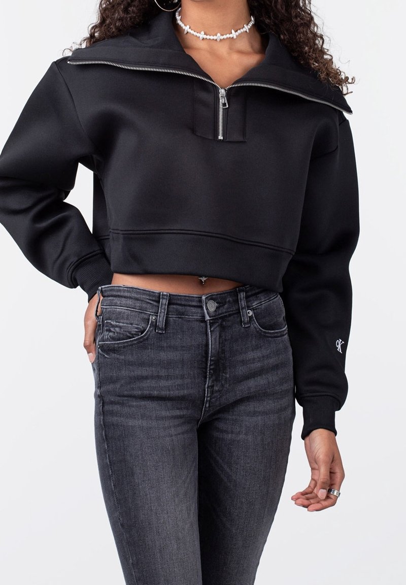 Femme portant un sweat-shirt court noir avec une fermeture éclair au quart et un jean taille haute gris foncé, accessoirisée d’un collier ras-de-cou en perles et de boucles d’oreilles créoles.