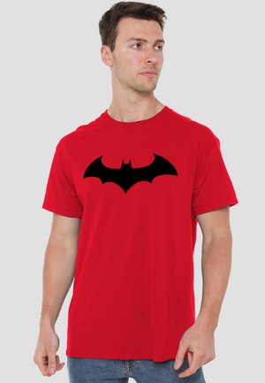Man wearing a red T-shirt with a large black bat emblem printed on the chest, looking to his right.