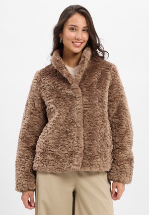 Jonge vrouw in een bruine fluffy nepbontjas, beige broeken en grote oorbellen, die glimlacht tegen een effen witte achtergrond.