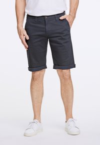 Shorts blu navy, lunghezza al ginocchio, realizzati in cotone con risvolti, dotati di una tasca frontale e chiusura a bottone. Indossati con scarpe da ginnastica bianche.