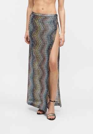 Nő, aki többszínű cikk-cakk mintás maxi szoknyát visel magas oldalsliccel és fekete nyitott orrú bokapántos szandált.