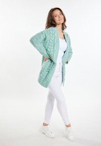 Turkos stickad cardigan med kabelmönster, öppen framsidan och oversized passform, kombinerad med vit topp och jeans, bärs med vita sneakers.