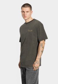 Oliwkowa bawełniana koszulka z krótkim rękawem, okrągłym dekoltem i małym, tłoczonym logo na piersi. Noszona z szarymi dżinsami.