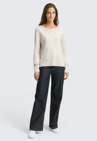 Top lungo beige chiaro a maniche lunghe con tessuto texturizzato e polsini a fantasia, abbinato a jeans a gamba larga blu scuro e sneakers bianche.