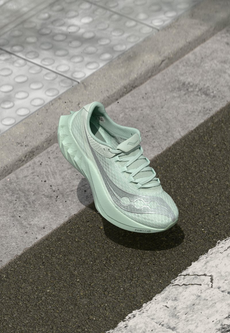 Heledroheline jooksutoss, millel on tekstureeritud võrkpinnas, peegeldavad hõbedased aktsendid ja eristuv, skulptuuriline tald. Paigutatud asfaldile.