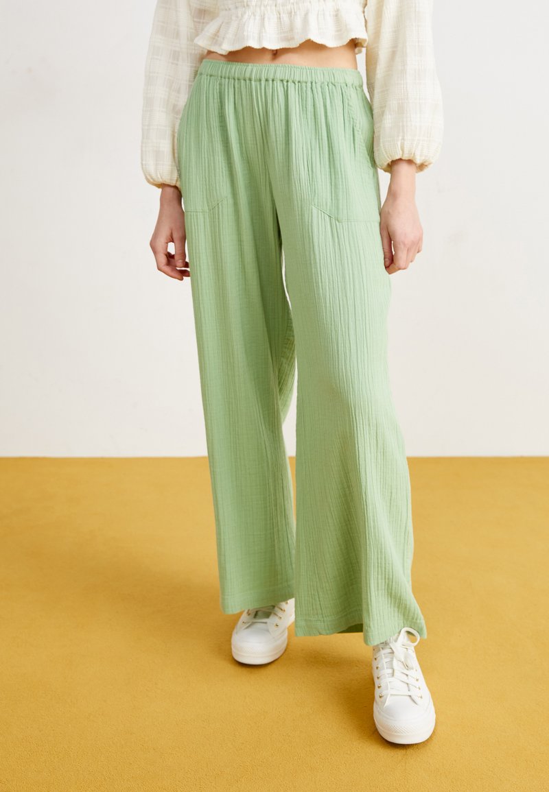 Light green, wide-leg pants with a textured fabric, featuring an elastic waistband and two front pockets, paired with white sneakers.