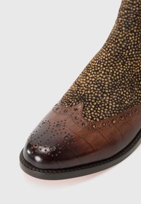 Botín de cuero marrón con un acabado brillante, que presenta un intrincado diseño de adornos y una parte superior texturizada y con un patrón en marrón y negro. Suela de perfil bajo.