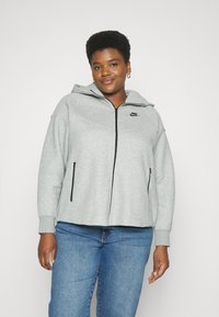 Pelēks nepiepildīts sporta jaka izgatavota no mīksta auduma ar kapuci, ar melniem akcentiem un Nike logo uz krūšu daļas. Priekšējās malās ir kabatas.