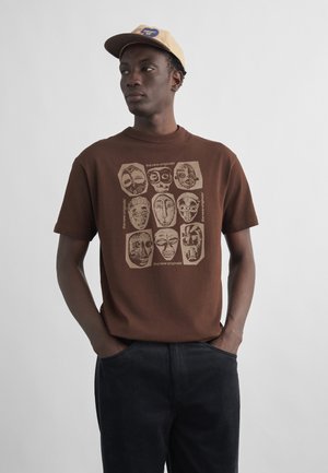 Mann iført brun T-skjorte med beige stammemønster i ansiktet og beige caps, stående med hendene i lommer mot ensfarget bakgrunn.