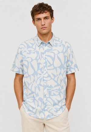 Mann trägt ein kurzärmliges Hemd mit blau-weißem abstraktem Muster und steht mit den Händen in den Taschen einer beigen Hose.