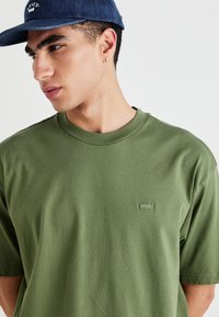Oliven grøn kortærmet T-shirt lavet af blødt bomuld, med et lille logo-patch på brystet og et klassisk rundhalset design.