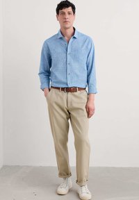Lysblå skjorte med knapper, beige bukser, brun vævet bælte og hvide sneakers. Skjorten har en brystlomme og lange ærmer.