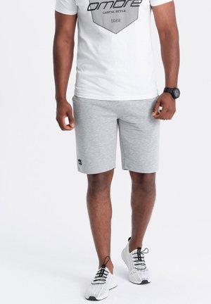 Shorts de sport gris avec une texture horizontale, associés à un t-shirt graphique blanc avec des lignes noires. Montre noire et chaussures de sport blanches.