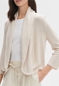 Beżowy kardigan z otwartym przodem, teksturowana dzianina, rękawy o długości 3/4, w połączeniu z białą bluzką w prążki i jasnymi spodniami.