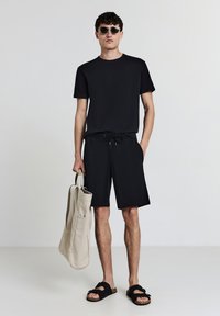 Schwarzes T-Shirt, schwarze Shorts mit Kordelzug, schwarze Sandalen und eine hellbeige Tragetasche. Glattes Material, schlichtes Design, lässiges Sommeroutfit.