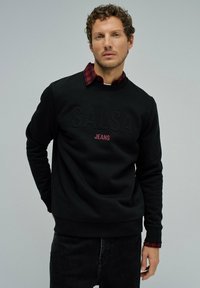 Sudadera negra con el logotipo en relieve de "SALSA" y el texto "JEANS" en rojo. Superpuesta a una camisa de cuadro rojo y negro, con cuello redondo.