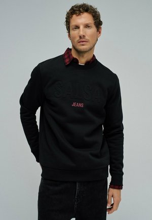 Sudadera negra con el logotipo en relieve de "SALSA" y el texto "JEANS" en rojo. Superpuesta a una camisa de cuadro rojo y negro, con cuello redondo.
