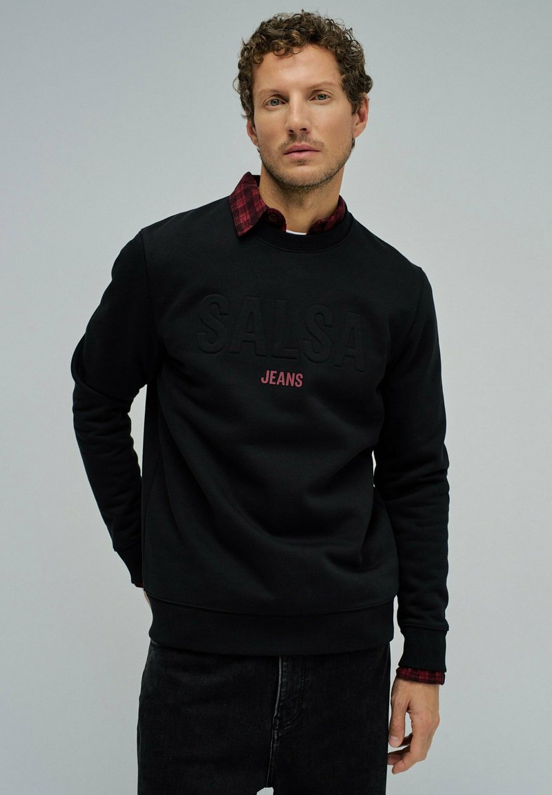 Sudadera negra con el logotipo en relieve de "SALSA" y el texto "JEANS" en rojo. Superpuesta a una camisa de cuadro rojo y negro, con cuello redondo.