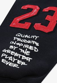 Černá látka s velkým červeným číslem "23" a bílým textem, který zní "KVALITNÍ PRODUKTY INSPIROVANÉ NEJLEPŠÍM HRÁČEM VŠECH DOB".