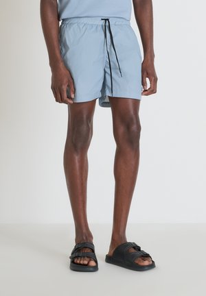 Pantaloncini da bagno azzurro chiaro con vita elasticizzata e cordoncino nero, abbinati a sandali neri scivolosi, che mostrano una texture liscia e leggera.