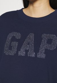 Námořnicky modrá mikina s texturovaným, třpytivým stříbrným logem "GAP". Látka je měkká a má kulatý výstřih s jednoduchým designem.