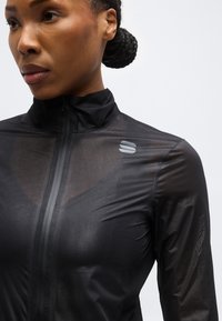 Γυναίκα με πλεξούδα σε κότσο που φορά εφαρμοστή μαύρη ζακέτα με φερμουάρ και μικρό αντανακλαστικό λογότυπο στον αριστερό ώμο, κοιτάζοντας προς τα αριστερά με φόντο λευκό.