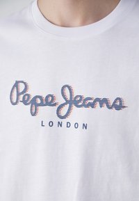 Baltos medvilnės marškinėliai su mėlynomis ir raudonomis juostelėmis: "Pepe Jeans" viršuje ir "LONDON" moderniu kreiviu šriftu. Apvalus iškirpimas, standartinė forma.