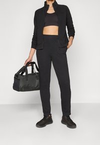 Chaqueta deportiva negra con cremallera, top corto negro, pantalones ajustados negros y zapatillas negras. Bolsa de deporte gris con detalles en negro sostenida en la mano.