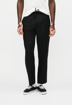 Isik, kes kannab musta pingutusnööriga pükse, valgeid sokke, musta-valgeid tossusid ja valget toppi, seisab ühevärvilise hele tausta ees.