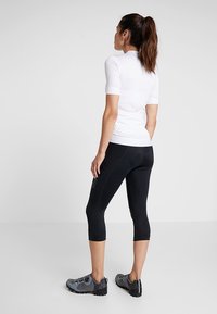 Top branco de manga curta, leggings pretos até à metade da panturrilha e sapatilhas desportivas cinzas. O tecido parece liso com um design ajustado. As leggings têm detalhe de logótipo.