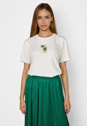 Frau trägt ein weißes T-Shirt mit einer kleinen Grafik einer grünen Pflanze und einen hoch taillierten, smaragdgrünen Faltenrock, steht vor einem schlichten Hintergrund.