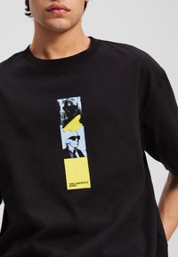 Svart bomull t-skjorte med et grafisk trykk med et farget rektangulært design, som viser en silhuett av en figur og tekst i gult.