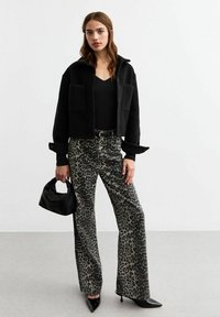 Chaqueta negra recortada con dos bolsillos frontales, combinada con pantalones de pierna ancha de estampado de leopardo. La modelo sostiene un pequeño bolso negro.