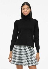Czarny sweter z wysokim kołnierzem noszony z spódnicą w wzór houndstooth w bieli i czerni. Oba elementy charakteryzują się luźnym krojem i gładką fakturą.