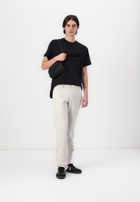 T-shirt noir à manches courtes avec impression en cursive, pantalons beige clair, baskets noires et un petit sac à bandoulière noir. Design simple et moderne.