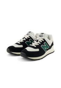 Une paire de baskets New Balance 574 noires et blanches avec des logos "N" verts, des lacets blancs et des semelles texturées sur fond blanc.