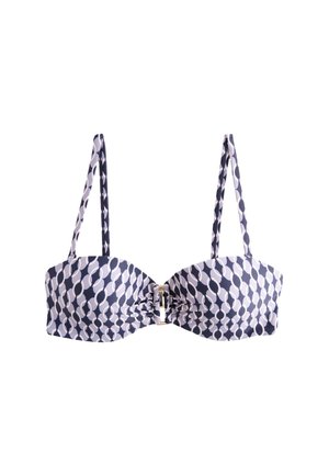 Kobieta z długimi ciemnymi włosami, ubrana w bezramiankowy, wzorzysty top od bikini, stoi na piaszczystej plaży z oceanem w tle.