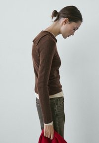 Woman with brown sweater and dark jeans looks down, holding a red coat in her right hand against a plain light background.