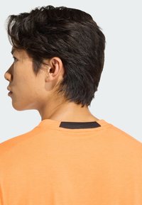 Camiseta deportiva naranja con cuello redondo. Presenta un detalle negro en la parte posterior del cuello. La tela parece suave y ligera.