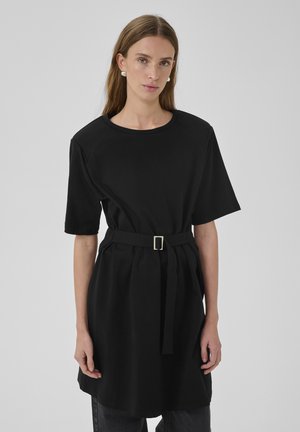 Frau mit langen Haaren, die ein schwarzes Tunika-Kleid mit Gürtel über dunkler Hose trägt, steht vor einem schlichten grauen Hintergrund.