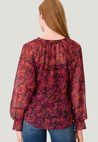 Haut transparent avec un motif paisley vibrant en rouge, violet et noir. Présente des manches longues, un col froncé et une coupe décontractée.