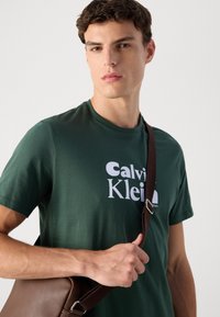 Young man wearing dark green Calvin Klein t-shirt and holding brown leather shoulder bag strap against plain light background.