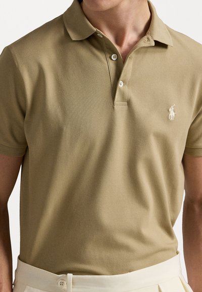 Khaki polo krekls ar klasisku apkakli, divu pogu pogu un izšūtu logotipu uz krūškurvja. Valkāts ar gaiša toņa biksēm.