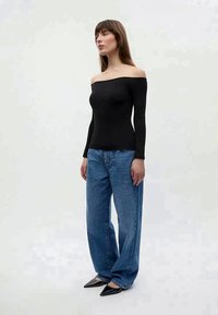 Black off-the-shoulder long-sleeve top paired with high-waisted blue jeans and pointed black heels, standing on a neutral background.
