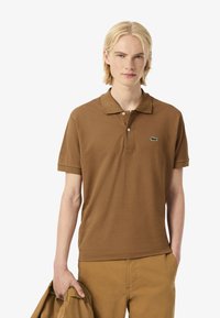 Hnědá bavlněná polokošile s krátkými rukávy, dvouknoflíková légou a zeleným logem na levé straně hrudi. Model drží hnědou bundu ve stejném odstínu.