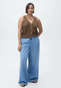 Brauner Strickweste mit Knopfverschluss, kombiniert mit hellblauen, weit geschnittenen Jeans. Die Sandalen haben einen schwarzen Riemen. Einfaches, lässiges Outfit.