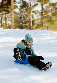Blå plastpulka med ett barn som sitter på den, iklädd en turkos jacka och en grå mössa. Snöigt bakgrund med träd. Barnet ler.