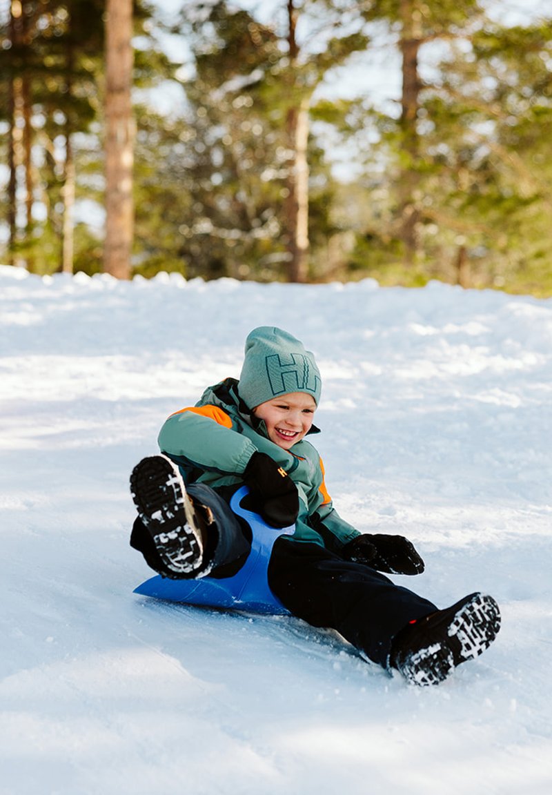 Blå plastpulka med ett barn som sitter på den, iklädd en turkos jacka och en grå mössa. Snöigt bakgrund med träd. Barnet ler.
