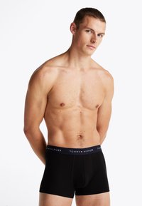 En mann uten skjorte, iført svarte Tommy Hilfiger boxer shorts, står med ryggen mot en enkel hvit bakgrunn og hendene bak ryggen.