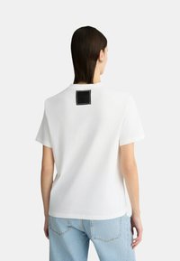 T-shirt bianca in cotone con maniche corte e scollo tondo, dotata di un'etichetta rettangolare nera cucita sul retro.